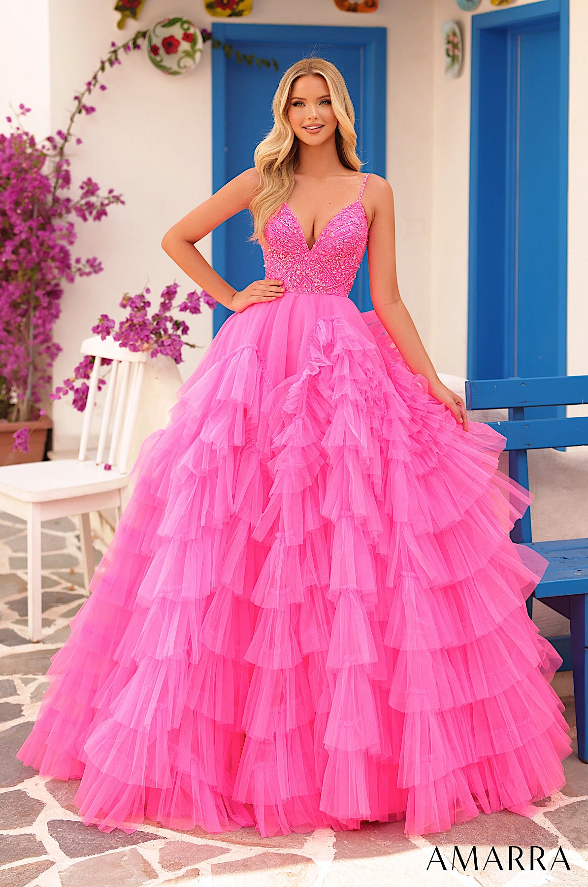 Woman in a pink ruffled dress standing in a decorative outdoor setting with flowers and blue chairs. ONLY AT THE BRIDAL COMPANY