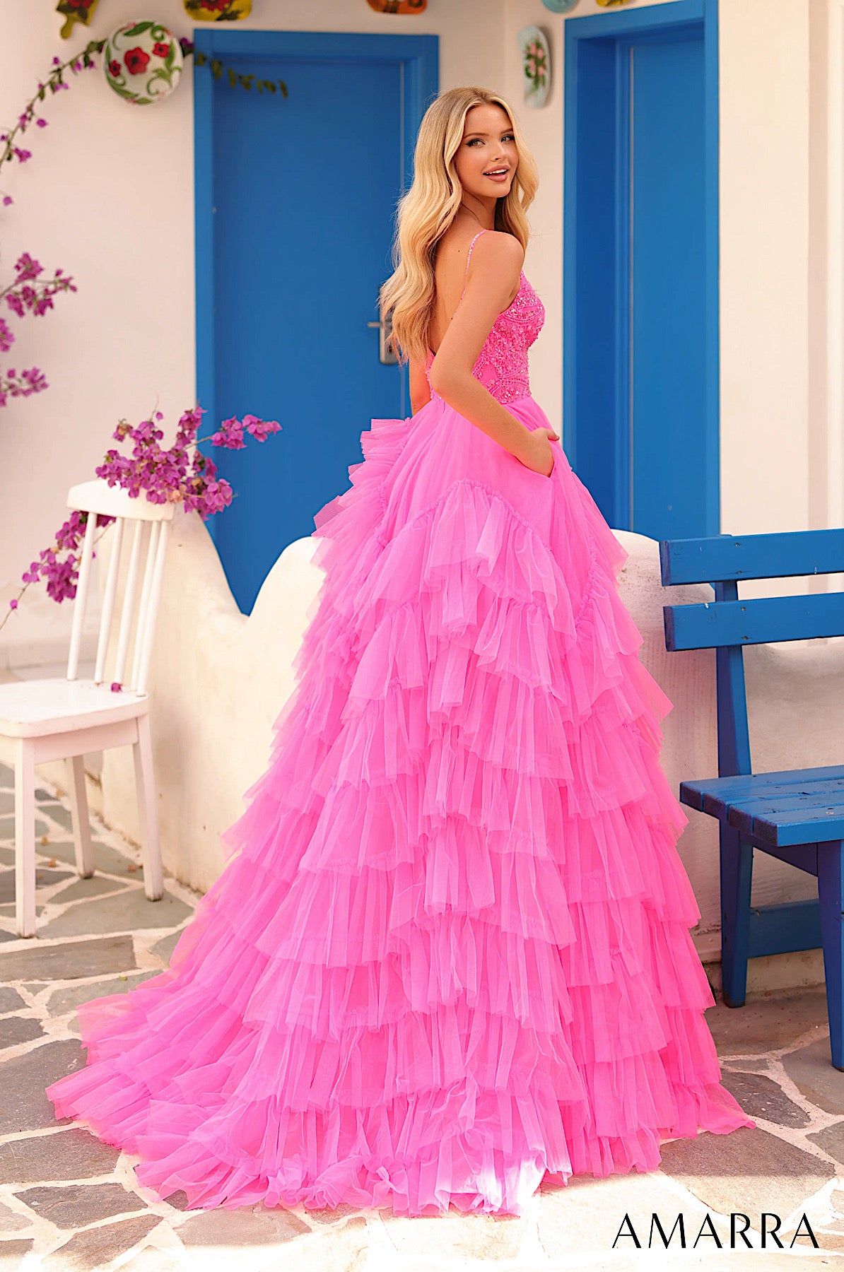 Woman in a pink ruffled dress standing in a decorative outdoor setting with blue doors and white walls. ONLY AT THE BRIDAL COMPANY