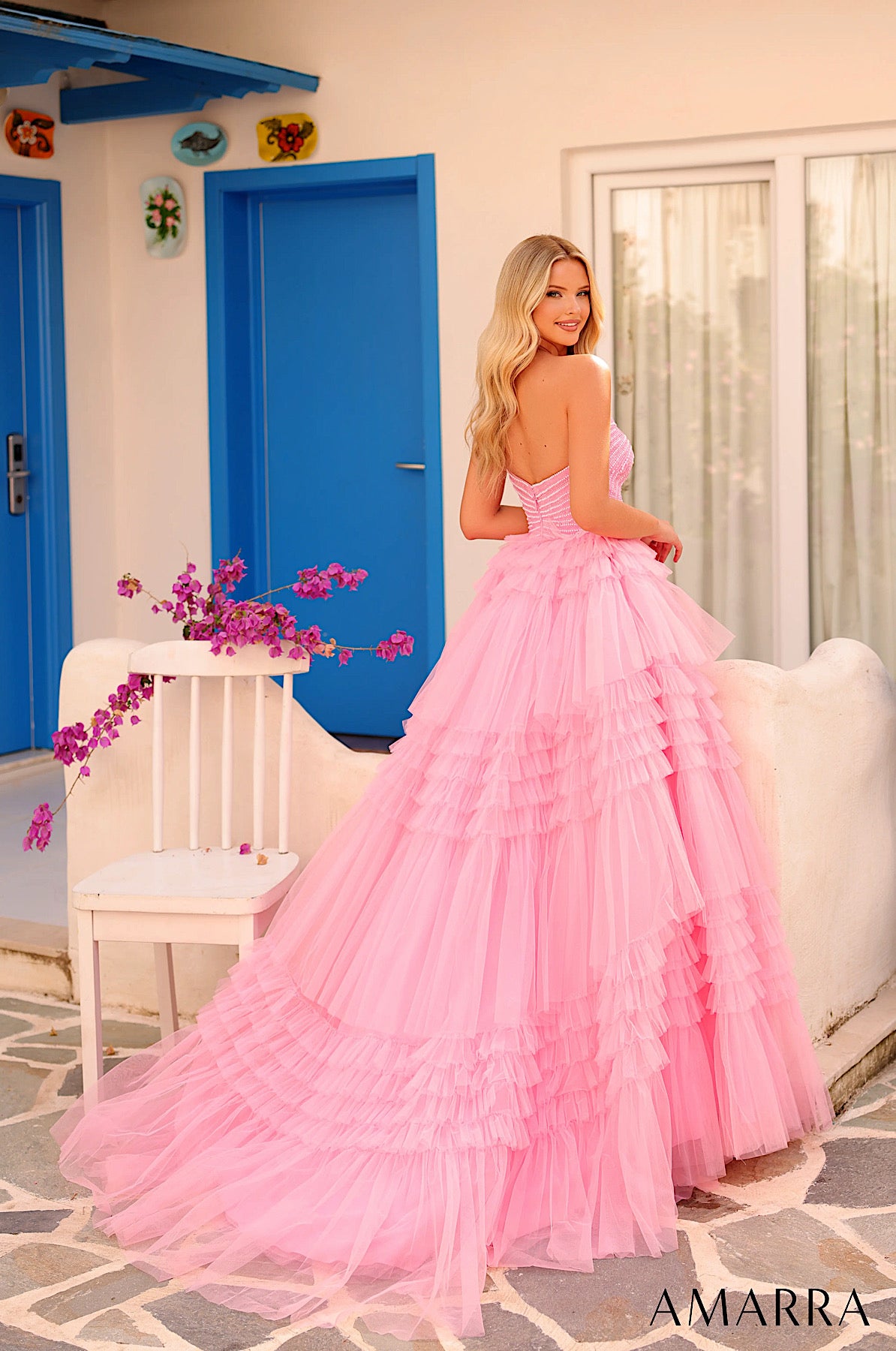 Woman in a pink dress standing in front of a blue door and white chair with purple flowers. ONLY AT THE BRIDAL COMPANY