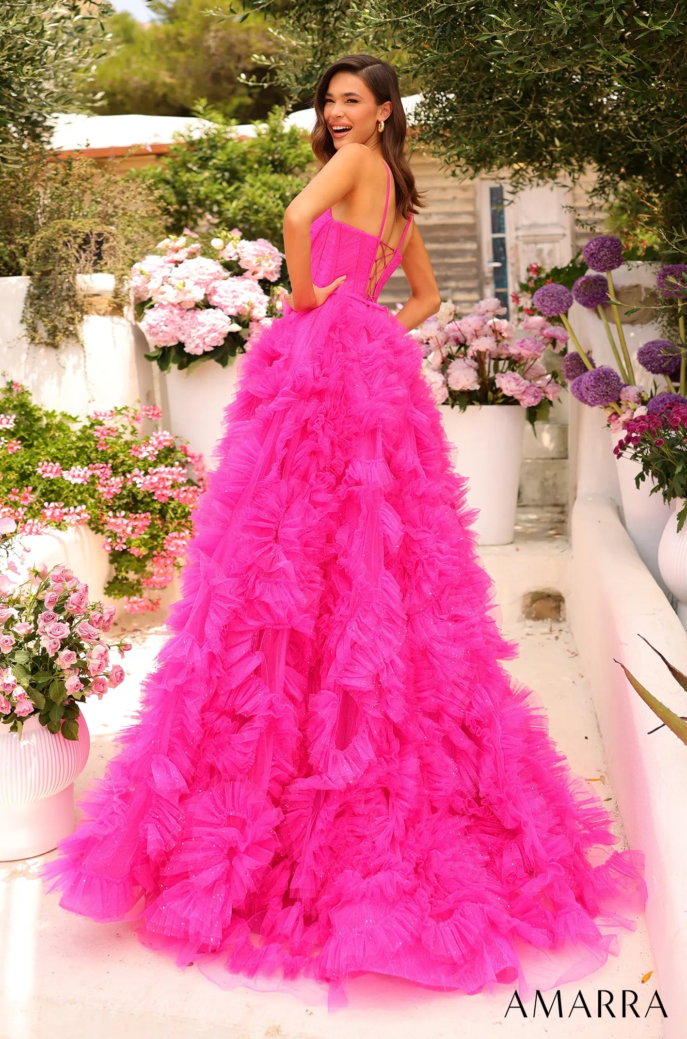Woman in a pink ruffled gown standing outdoors with flowers and white structures in the background
