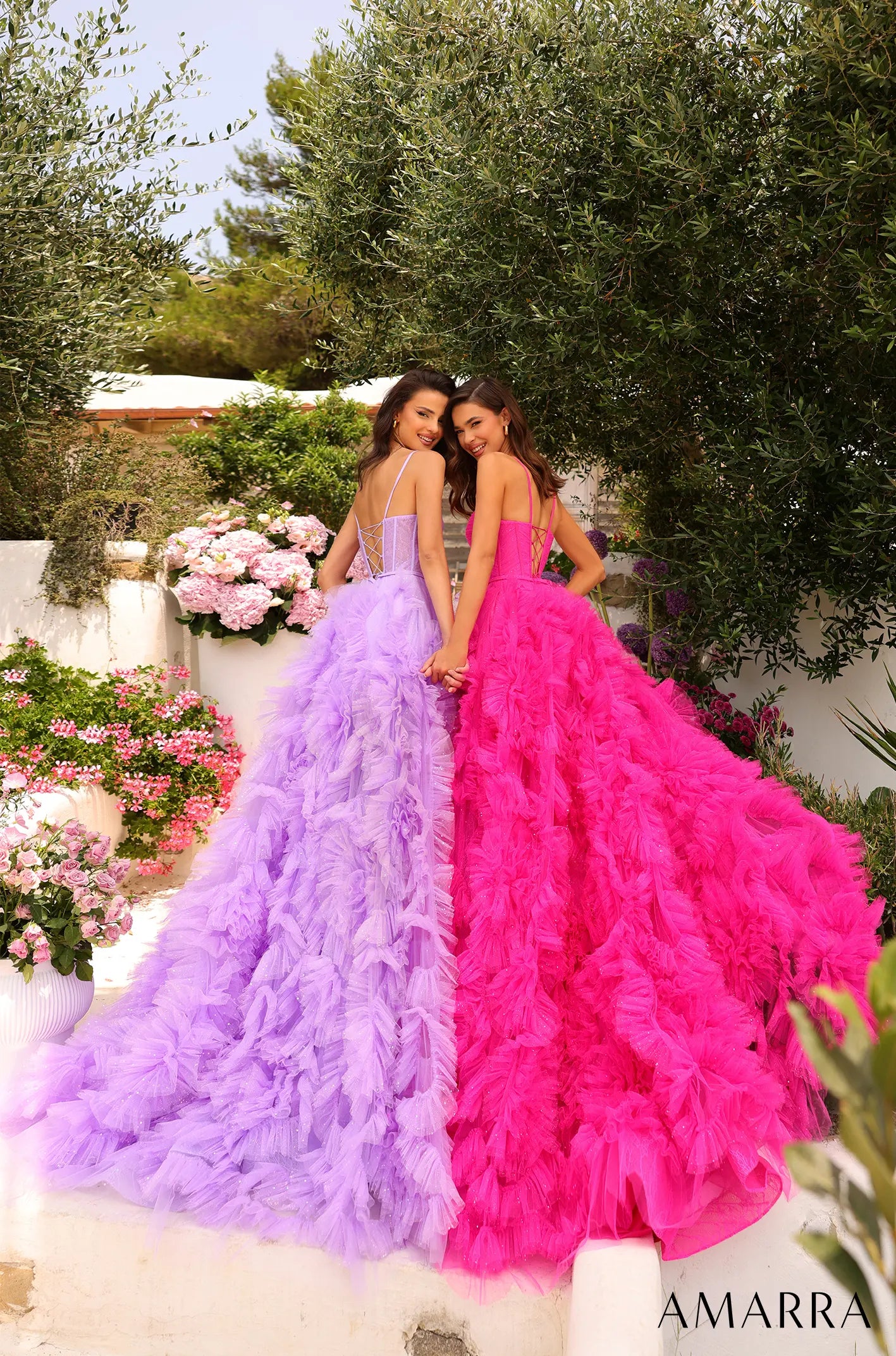 Two women in colorful ruffled dresses standing outdoors with flowers and greenery.