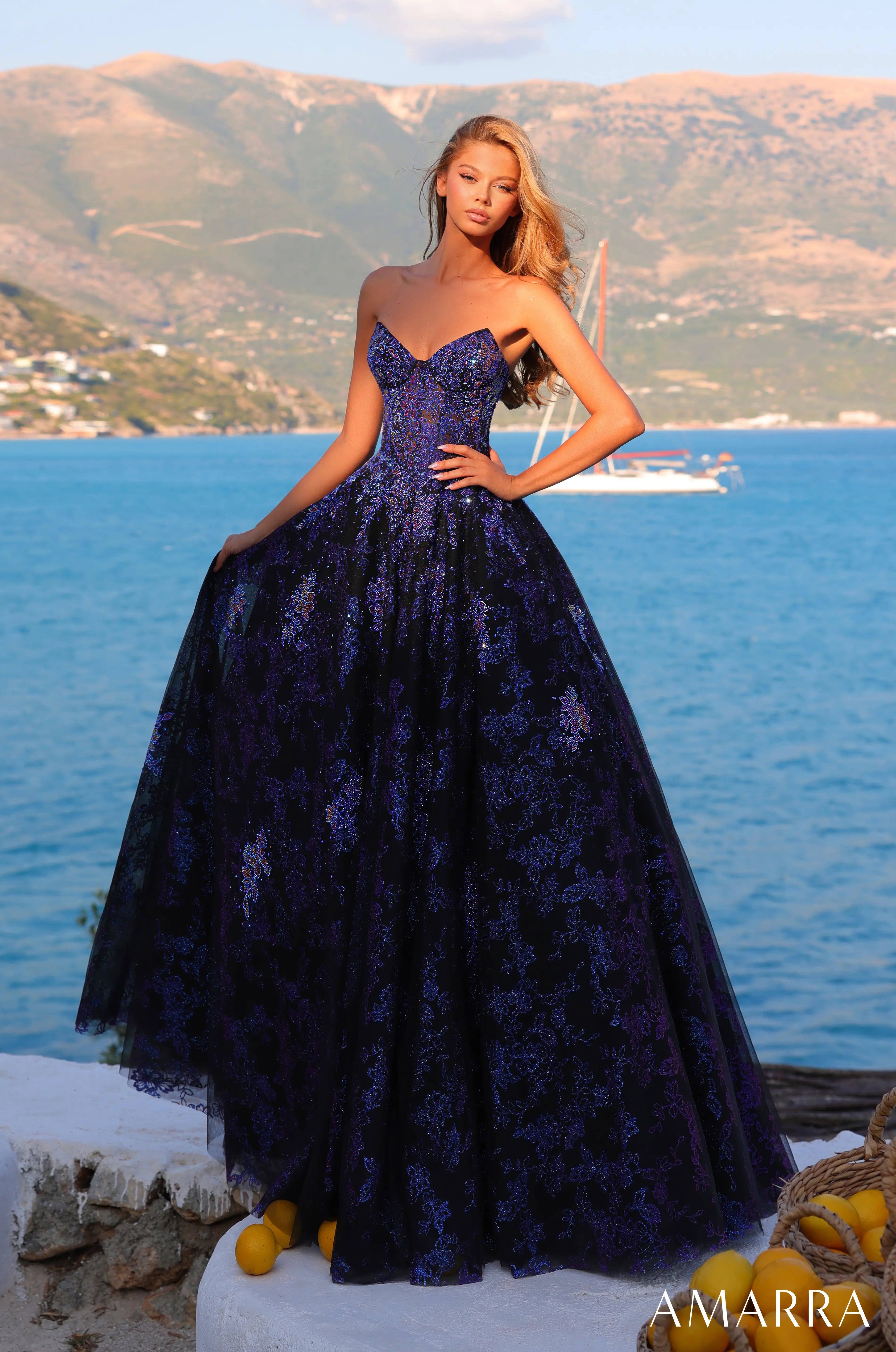 Woman in a dark blue evening gown standing by a scenic waterfront with mountains and water.