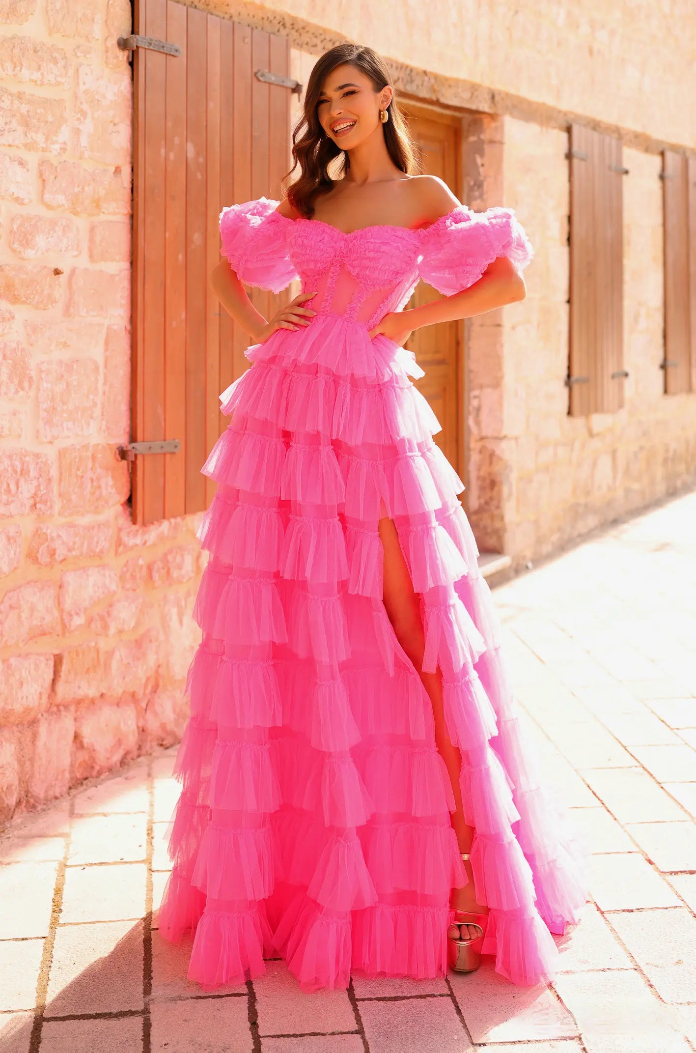 Woman wearing a pink ruffled dress standing in front of a stone wall. ONLY AT THE BRIDAL COMPANY