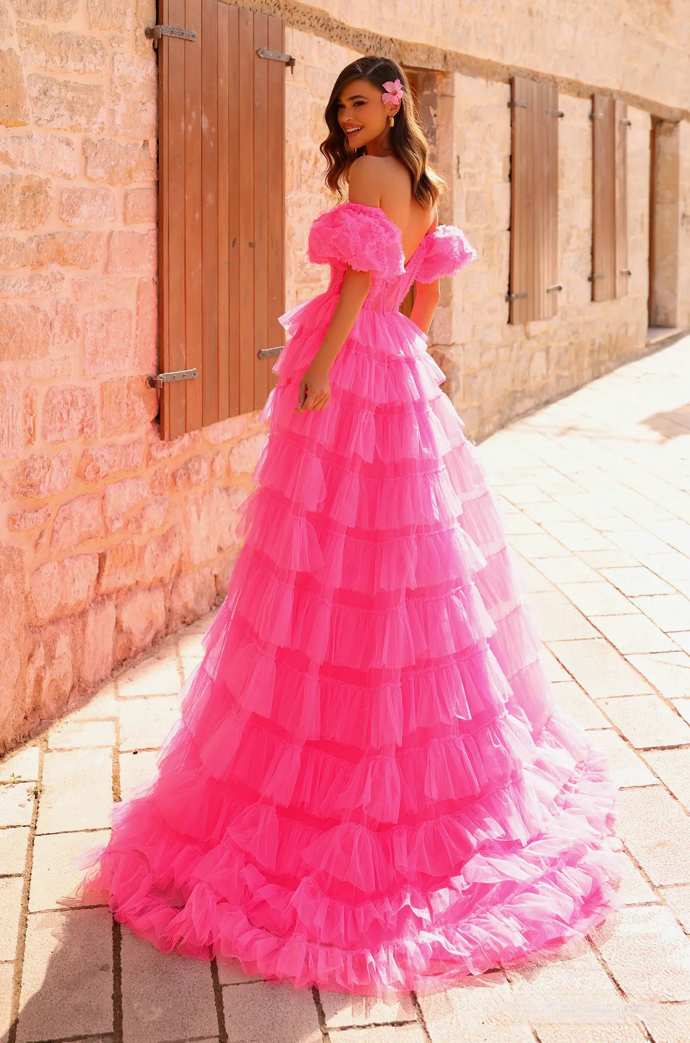 Woman in a pink off-shoulder dress standing against a brick wall. ONLY AT THE BRIDAL COMPANY