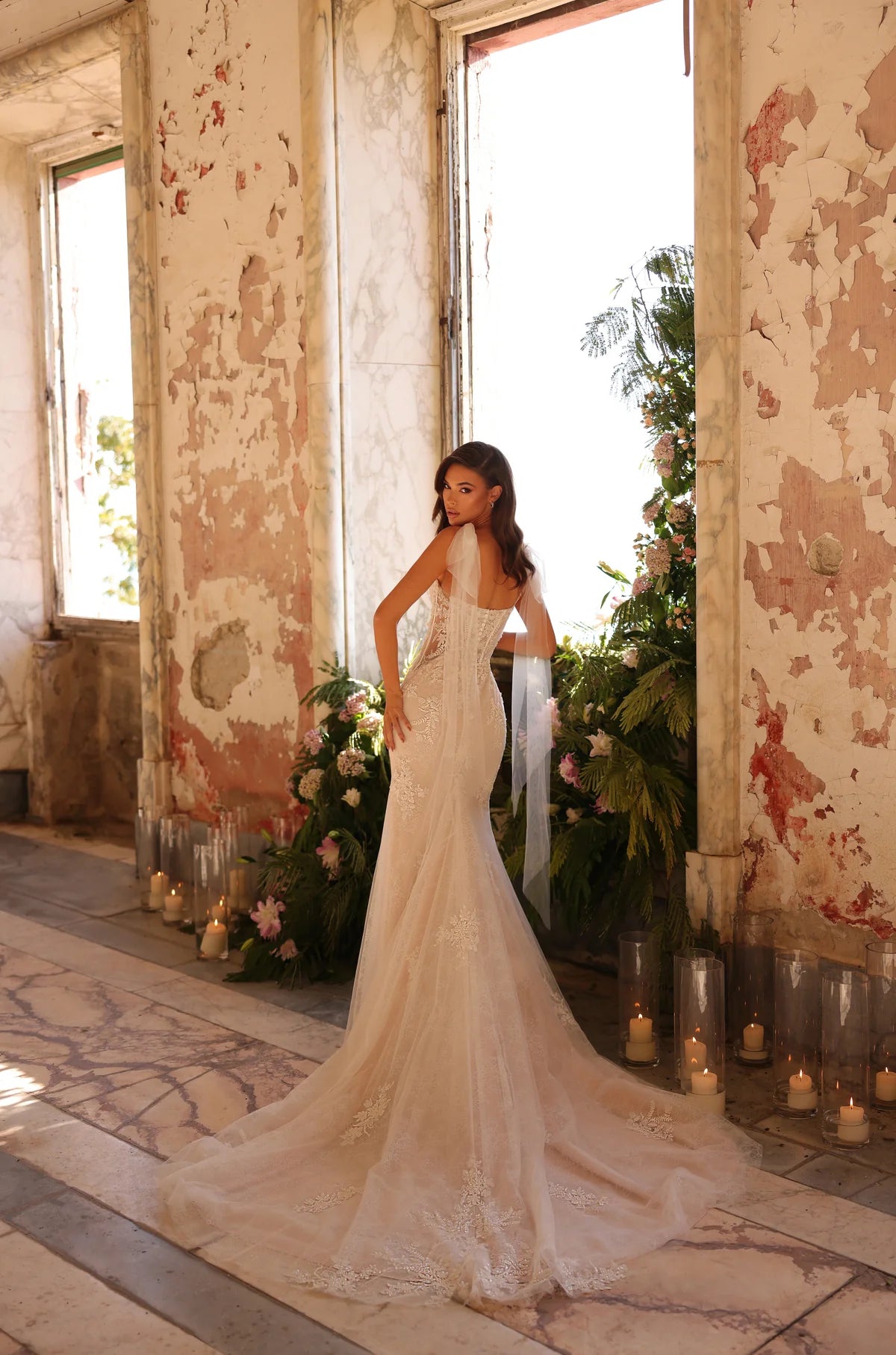 Woman in a white wedding dress standing in an elegant interior setting with candles and floral arrangements.