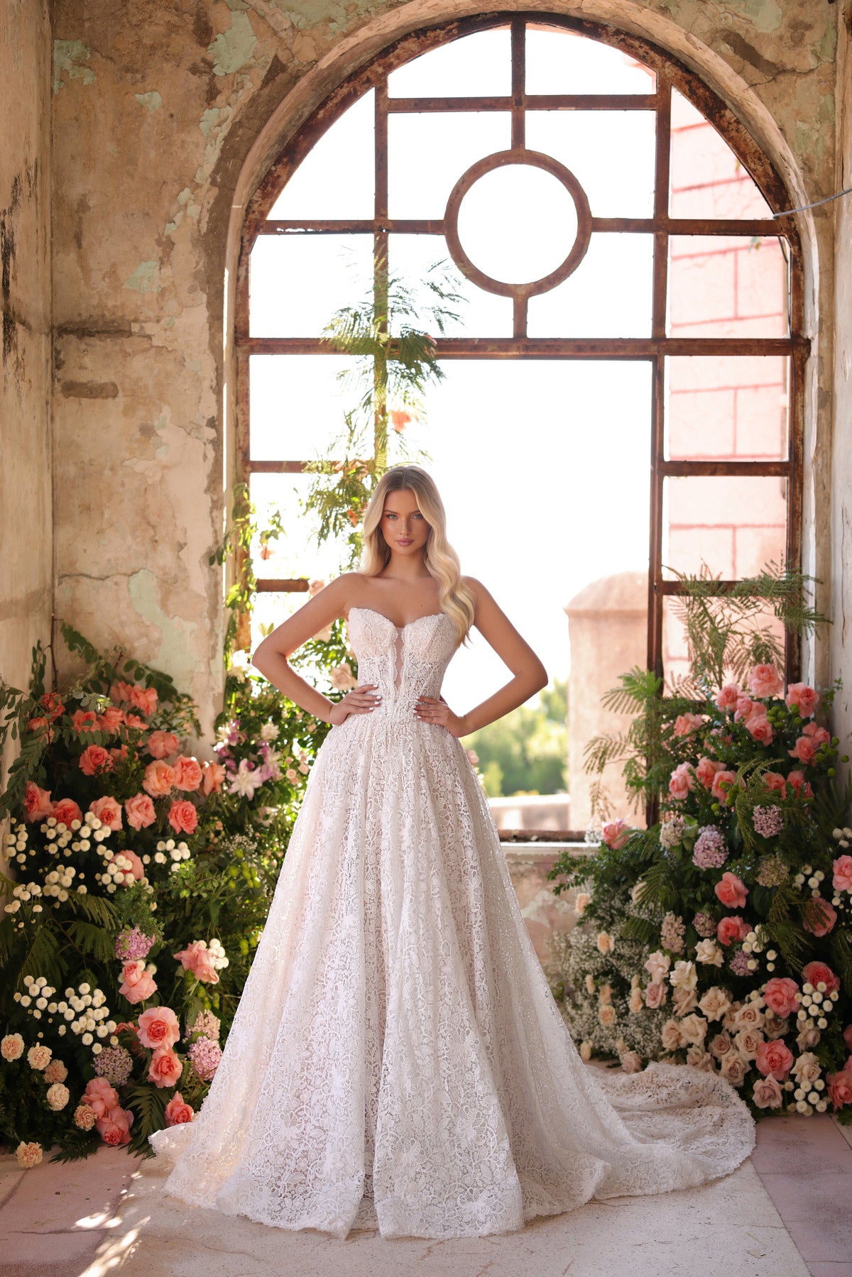 Woman in a white wedding dress standing in front of a large arched window with floral decorations.
