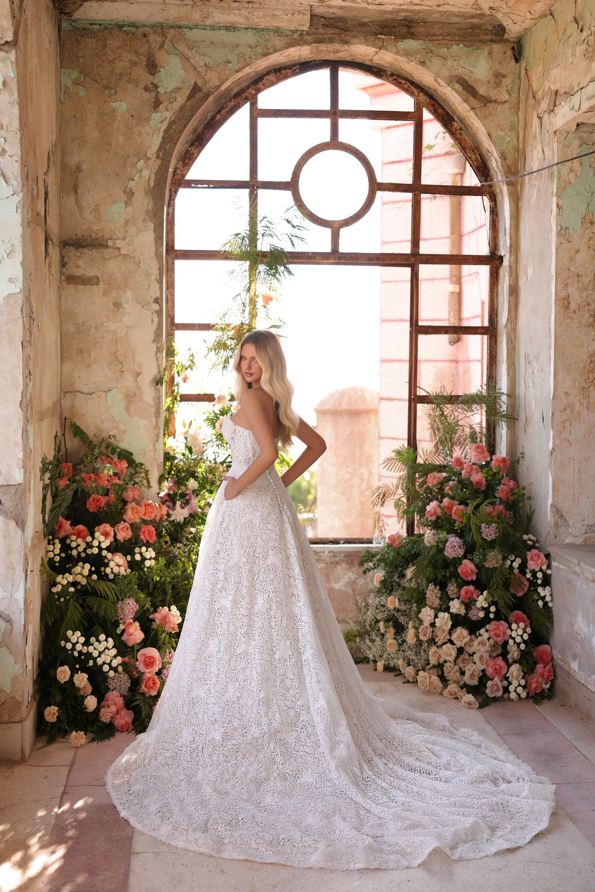 Woman in a white lace wedding dress standing in a decorative interior with floral arrangements and large windows.