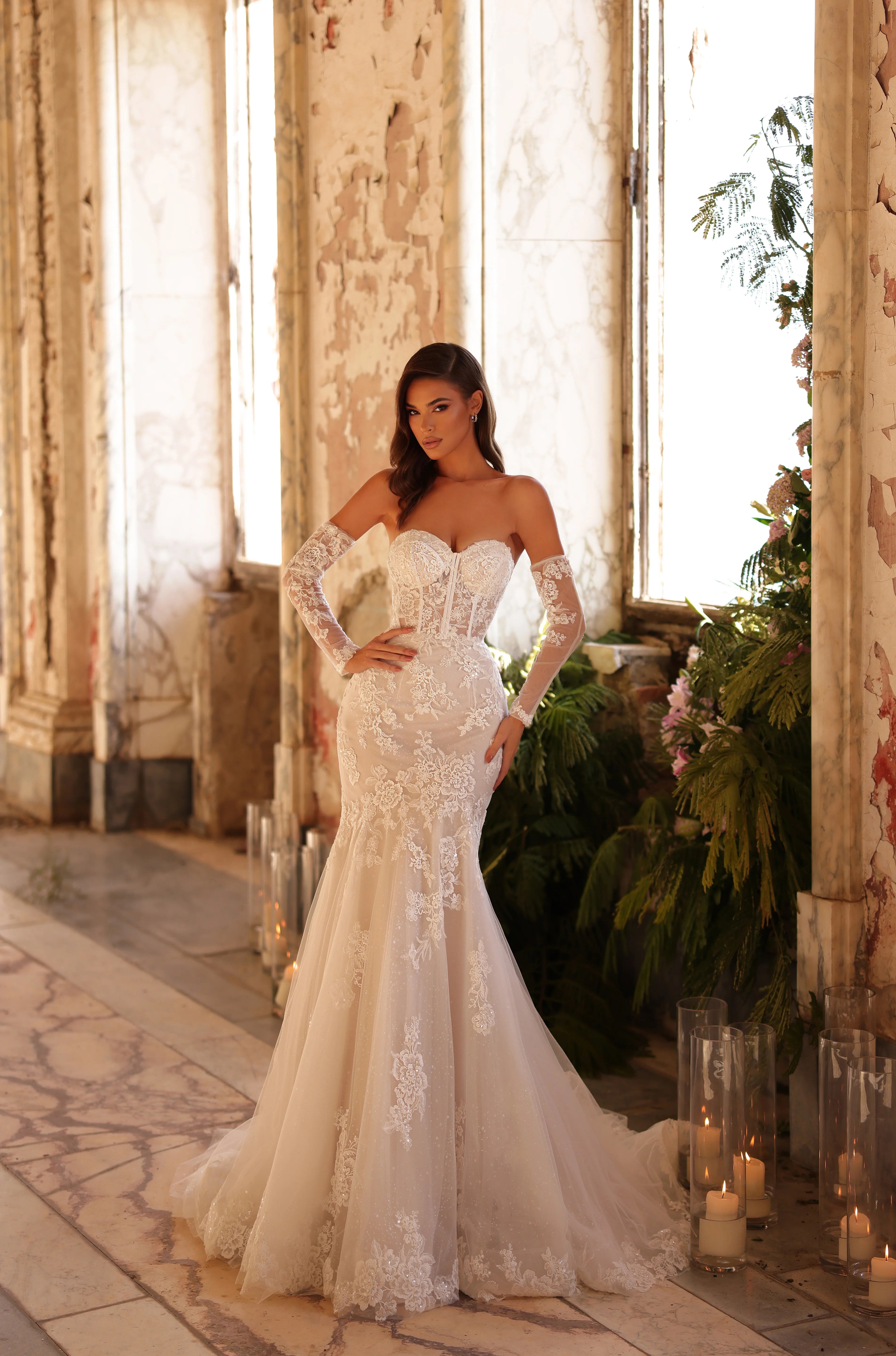 Woman in a white lace wedding dress standing in an elegant indoor setting with large windows and decorative elements.