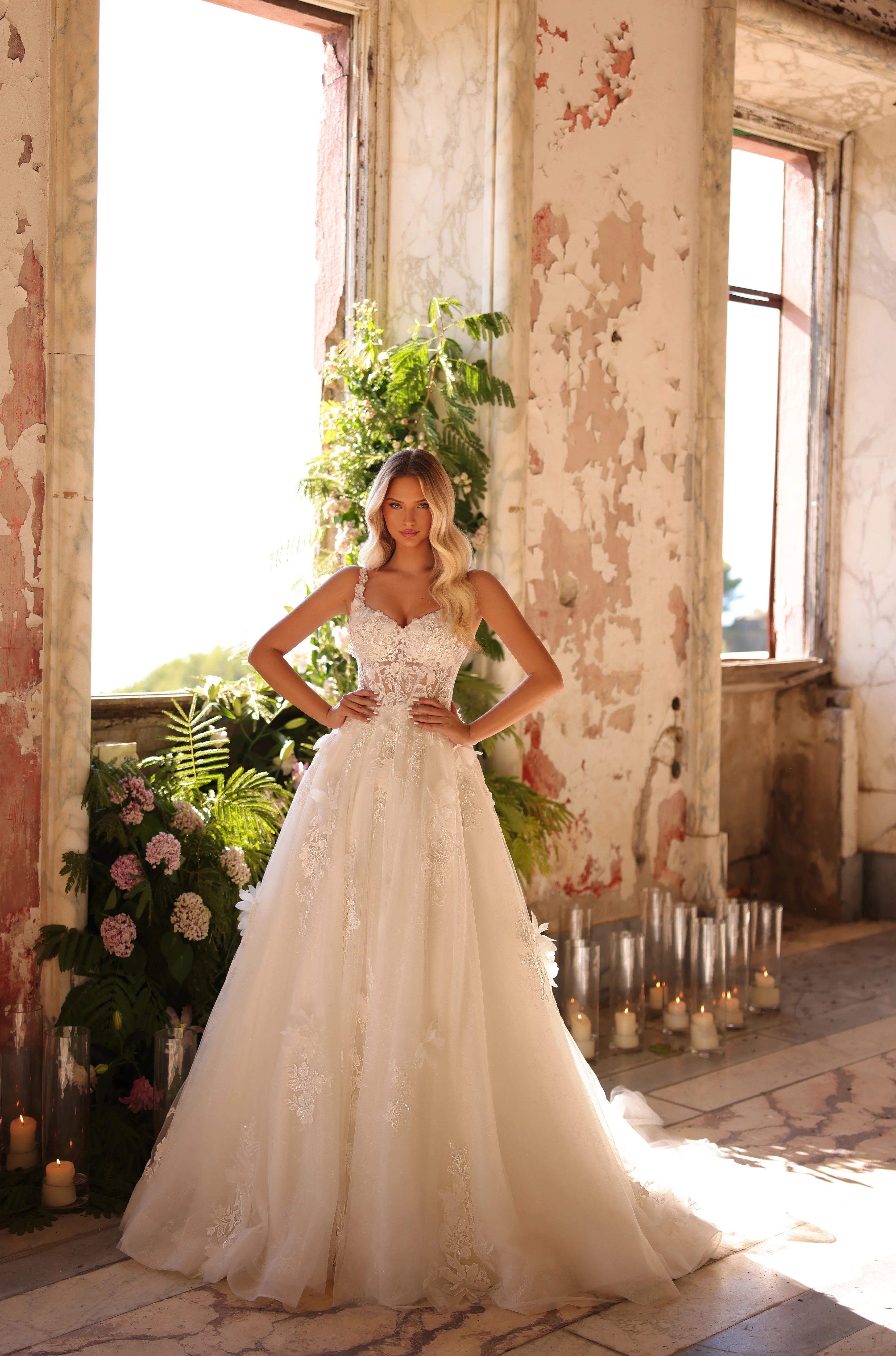 Woman in a white wedding dress standing in a rustic indoor setting with large windows and floral decorations.