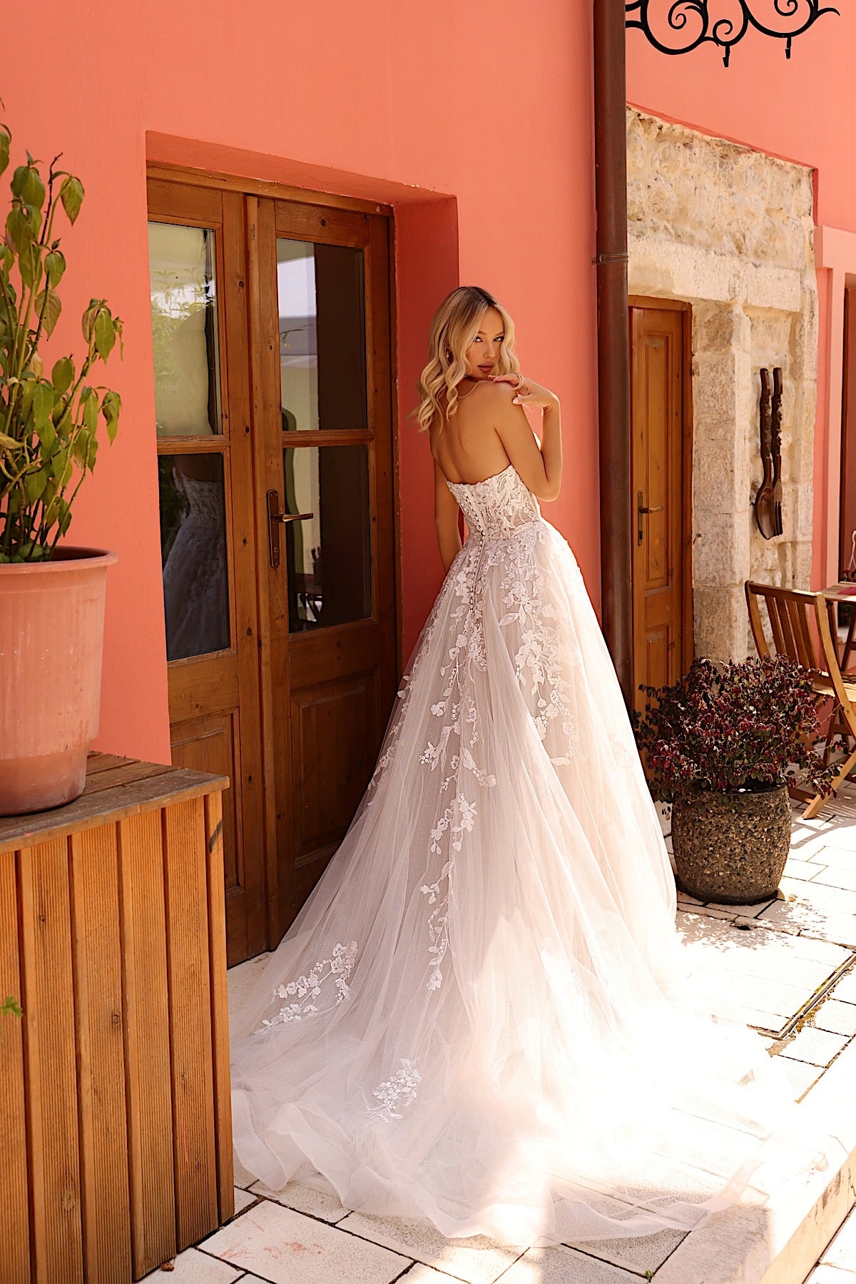 Woman in a white lace wedding dress standing in a sunlit room with pink walls and wooden furniture.