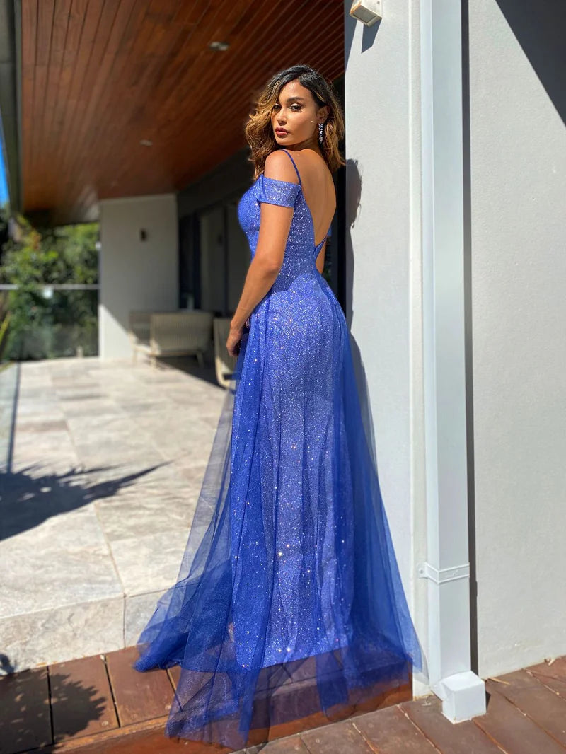 A woman wearing a royal blue off-the-shoulder evening gown with a sparkling lace bodice and a flowing tulle skirt, posing outdoors against a modern building wall. the bridal company