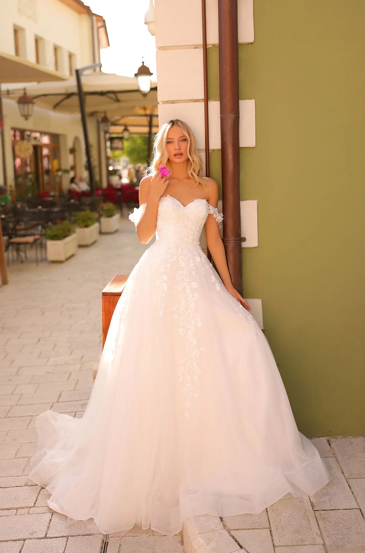 Woman in a white wedding dress standing on a street with buildings and plants in the background, with THE BRIDAL COMPANY branding.