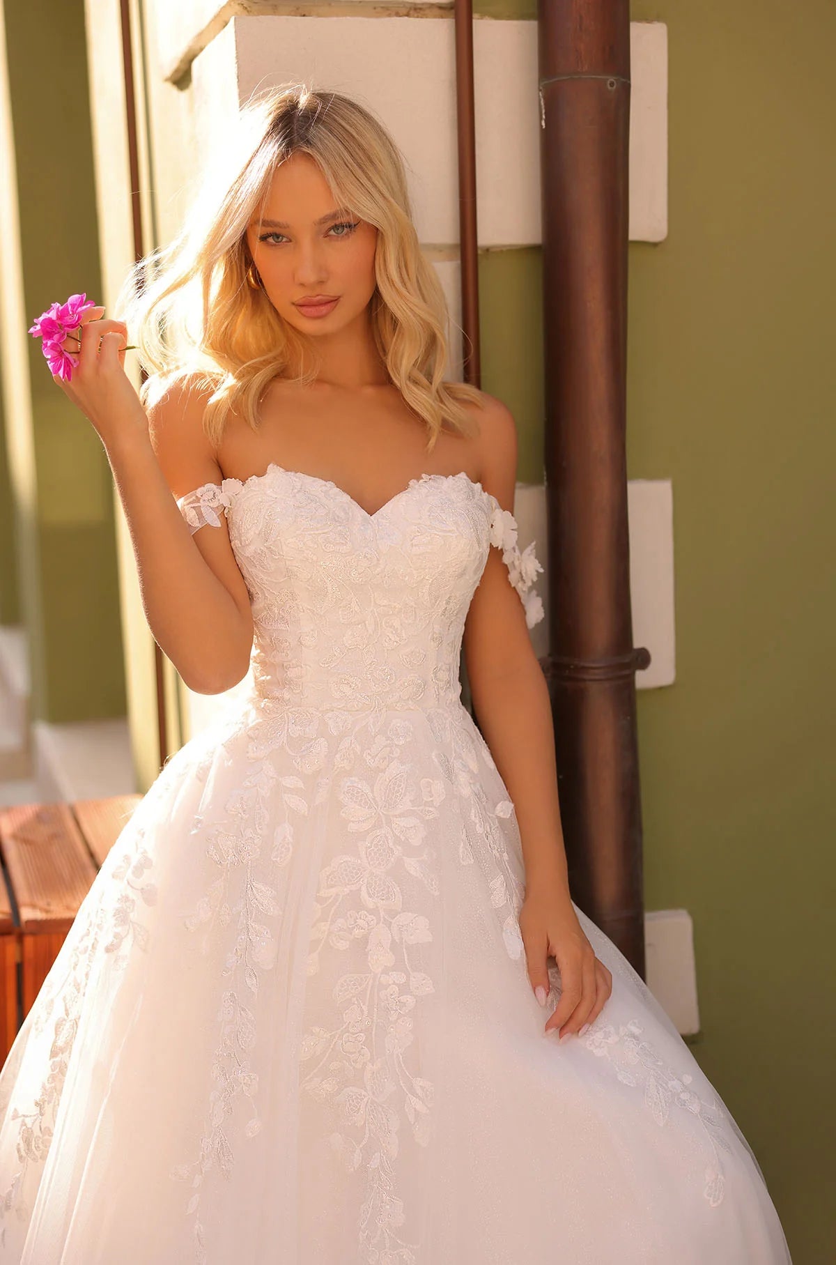Woman in a white wedding dress holding a pink flower against a green wall, with THE BRIDAL COMPANY branding.
