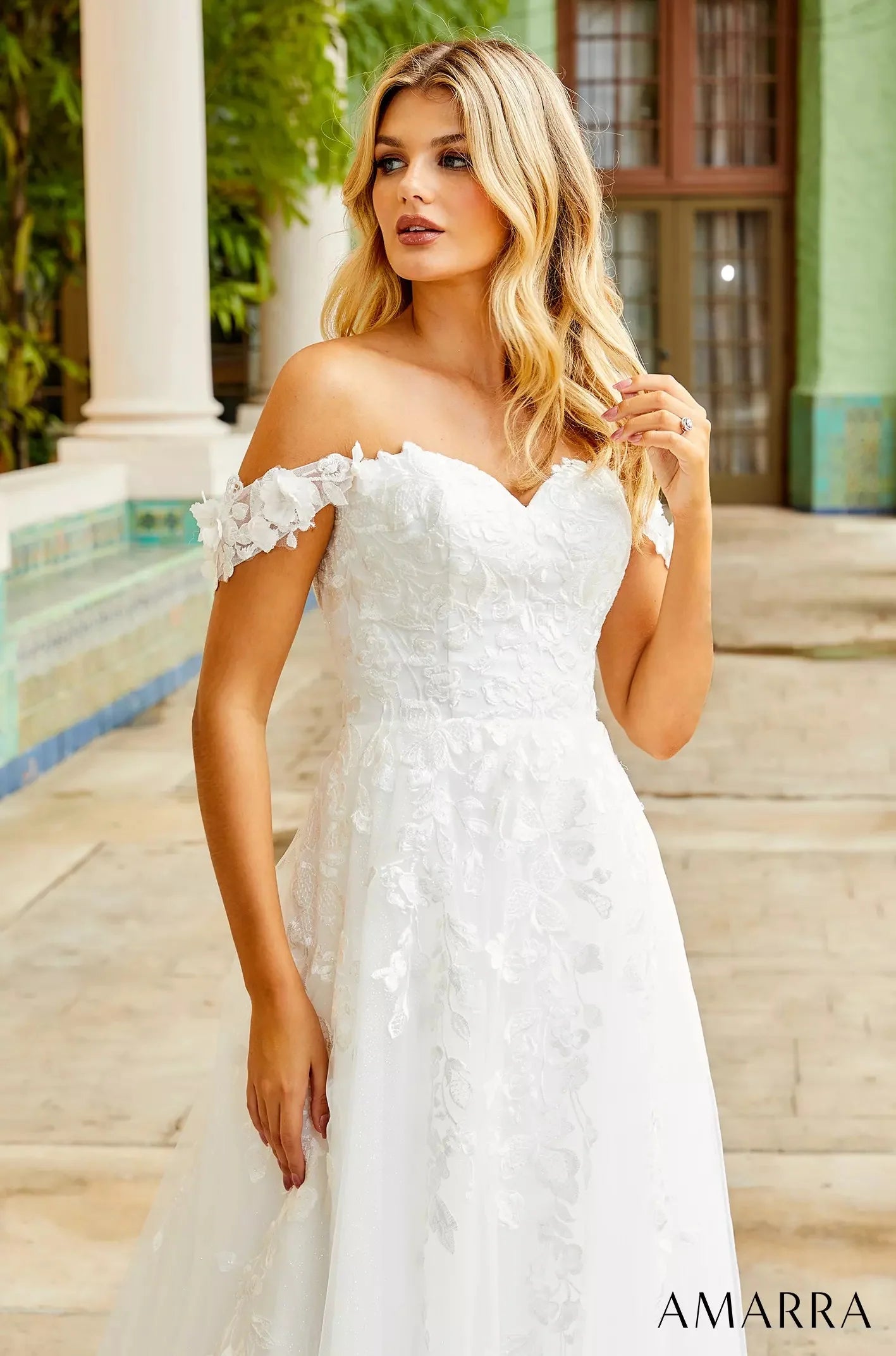 Woman wearing a white off-shoulder dress with floral details in an outdoor setting, with THE BRIDAL COMPANY branding.