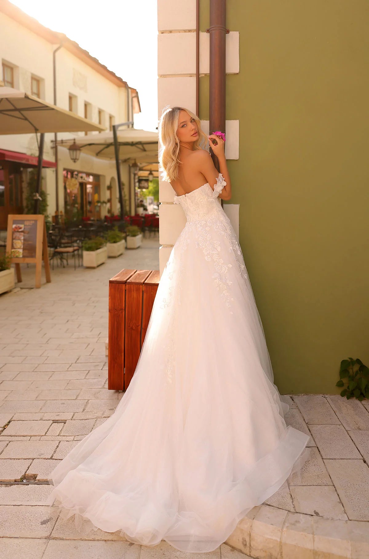 Woman in a white wedding dress standing on a street with buildings and plants in the background, with THE BRIDAL COMPANY branding.