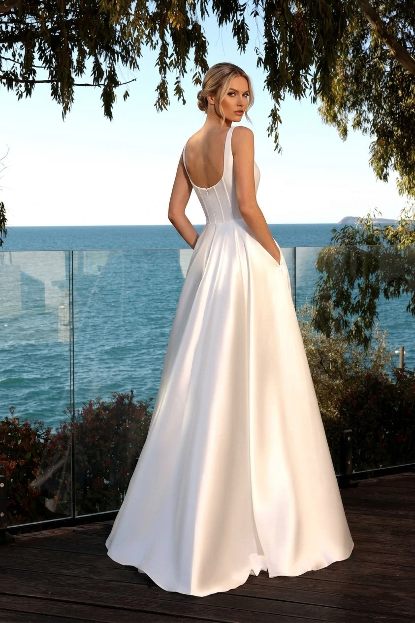 A woman wearing a white sleeveless wedding gown stands on a balcony overlooking the ocean, photographed from the back. The dress has a fitted bodice, sheer straps, a low open back, and a full flowing skirt available at the bridal company gold coast australia