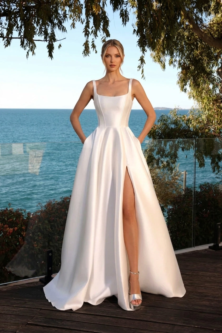 A woman models a floor-length white satin gown with thin straps, a fitted bodice, and a high thigh slit, standing on a wooden deck with the ocean and trees in the background available at the bridal company gold coast australia