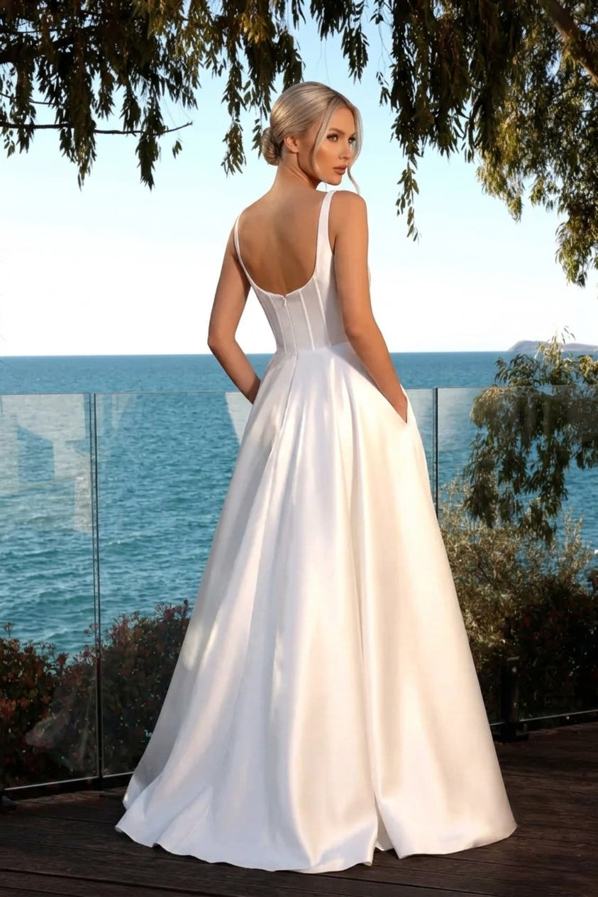 A woman wearing a white satin A-line wedding dress with a low open back and pockets, standing on a wooden deck with the ocean and trees in the background available at the bridal company gold coast australia