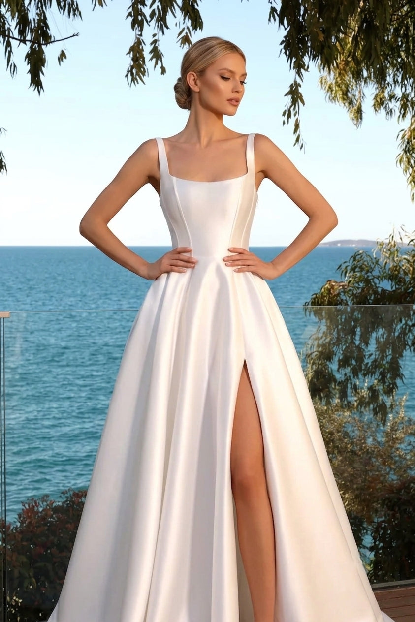 A woman modeling a white satin A-line evening gown with a thigh-high front slit, standing outdoors with the ocean and trees in the background available at the bridal company gold coast australia