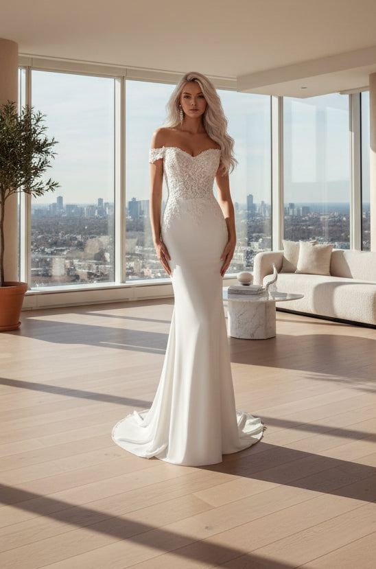 A woman modeling an off-the-shoulder white wedding gown with a fitted lace bodice and a floor-length satin skirt, posed against a dark blue spotlighted studio background.